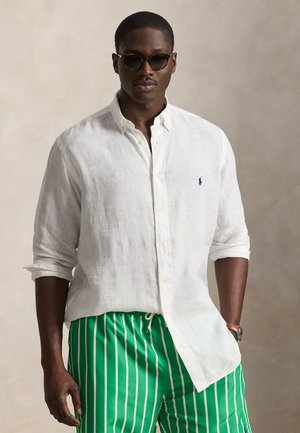 Vyras, dėvintis susuktą baltą marškinių su sagomis rankovę, žalias ir baltas dryžuotas šortus, akinius nuo saulės ir laikrodį ant riešo neutraliame fone.