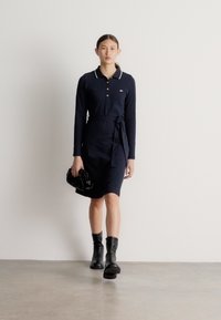 Tumši zils polo kleitas modelis ar garām piedurknēm un jostu viduklī, kontrastējoša apkakle, valkātas kopā ar melniem potīšu zābakiem un mazu melnu rokassomu.