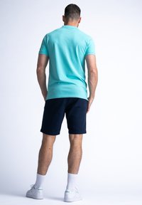 Tyrkysové polo tričko s krátkým rukávem v kombinaci s navy kraťasy. Model otočený zády, viditelná bavlněná struktura a bílé tenisky se zelenými akcenty.