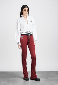 Bela zip-up jakna z ribastimi manšetami, v kombinaciji z maroon razširjenimi hlačami z bočnimi črtami in izvezenim logotipom blizu pasu. Črne čevlje.