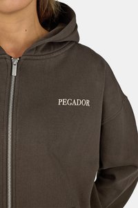 Barna, cipzáros kapucnis pulóver, puha anyagból készült, kapucnival, bordás mandzsettákkal és a "PEGADOR" szóval krémszínben az elején.