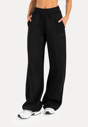 Persona vistiendo pantalones de chándal negros de pierna ancha con costuras verticales y zapatillas blancas, con las manos en los bolsillos, sobre un fondo claro liso.