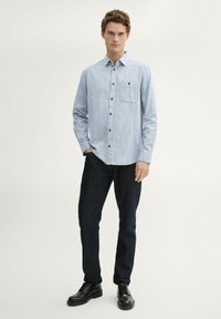 Lichtblauwe langemouwen overhemd met een button-down kraag, een borstzakje en donkere knopen, gecombineerd met donkerblauwe jeans en zwarte veterschoenen.