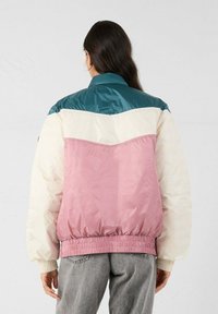 Persone che indossa una giacca a pannelli colorati con sezioni in teal, crema e rosa, abbinata a jeans grigi, vista da dietro su uno sfondo neutro.