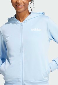 Light blue zip-up hoodie made of smooth fabric with a hood. Features white Adidas logo on the chest and elastic cuffs at the sleeves.