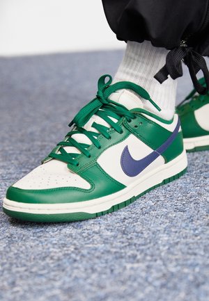 Baskets basses en cuir vert et blanc, logo Nike violet, lacets verts, portées avec des chaussettes blanches et un pantalon noir sur un sol moquetté bleu.