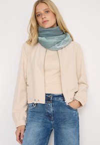 Chaqueta beige corta con puños elásticos, top beige claro y vaqueros azules. Una bufanda de color teal texturizada en diseño circular alrededor del cuello.