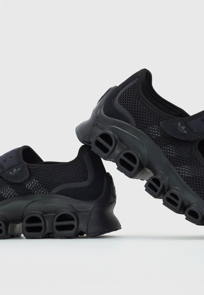 Chaussures de sport noires avec un dessus en mesh, un bout arrondi et un design de semelle distinct en forme de bulle avec plusieurs trous de ventilation. Sangle ajustable.