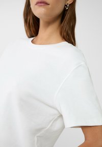 T-shirt bianco in cotone con maniche corte, scollo rotondo e superficie liscia. Presenta un design minimale senza motivi visibili o dettagli in metallo.