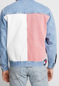 Persona che indossa una giacca di jeans color block con pannelli bianchi, rosa e azzurri e un logo Tommy Jeans vicino all'orlo.