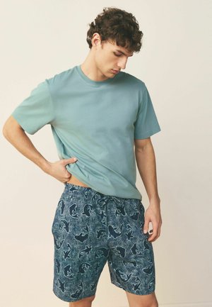 Fiatal férfi türkiz pólóban és sötétkék, cápa mintás rövidnadrágban, a rövidnadrág derékrészét igazítja miközben lefelé néz.