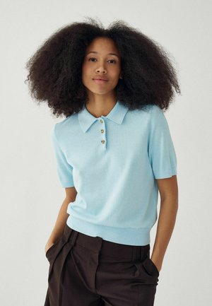 Noor naine volüümika loomuliku soenguga, kandes helesinist lühikese varrukaga polosärki ja tumedaid pükse, seisab käed taskutes.