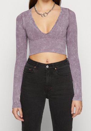 Frau trägt ein langärmliges, geripptes lila Croptop mit tiefem V-Ausschnitt, schwarze High-Waist-Jeans und eine dicke silberne Kettencollier.