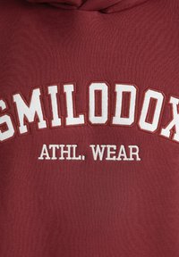 Sudadera burdeos con texto bordado en blanco que dice "SMILODOX ATHL. WEAR." Textura suave con un diseño atlético estructurado.