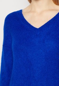 Pull à col en V fabriqué en tissu tricoté bleu doux et duveteux, avec des manches longues et une finition texturée.