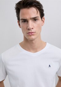 T-shirt bianca in cotone con scollo a V, con un piccolo emblema navy di teschio e ossa incrociate sul petto a sinistra. Tessuto liscio e vestibilità regolare.