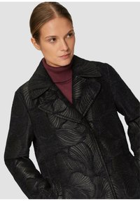 Manteau noir à motifs avec un design texturé, grand col et poches avant, porté avec un col roulé bordeaux foncé en dessous.