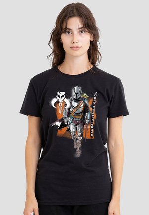Woman wearing black t-shirt featuring Mandalorian in armor and Grogu on his shoulder with orange graphic and text "Mandalorian & Grogu."