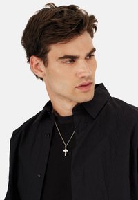 Sort skjorte med knapper og struktureret finish, lagdelt over en sort t-shirt. En sølvkorshalskæde er synlig ved halsen.