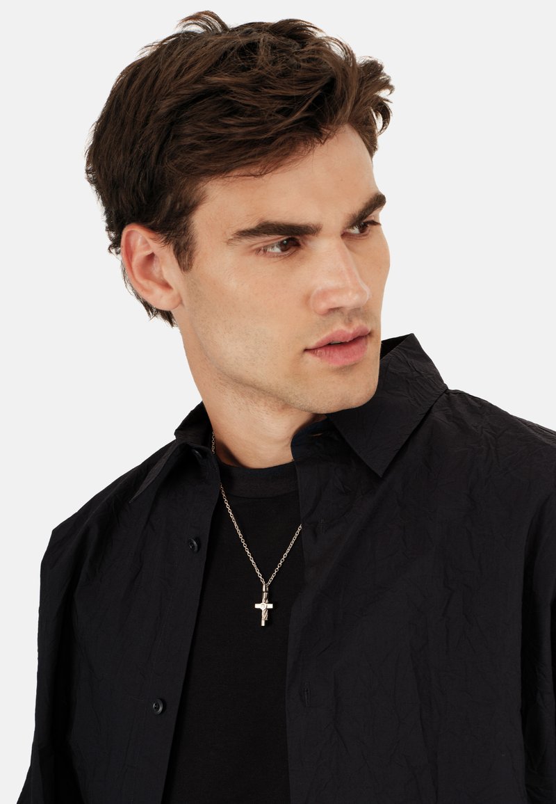 Sort skjorte med knapper og struktureret finish, lagdelt over en sort t-shirt. En sølvkorshalskæde er synlig ved halsen.