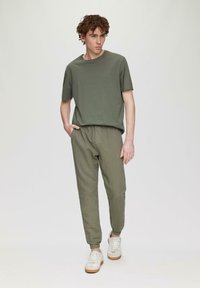 Oliwkowa koszulka z krótkim rękawem, w połączeniu z jasnooliwkowymi spodniami taperowanymi i białymi trampkami, stojąca na jasnym tle.