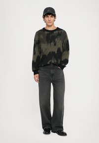 Pull en tricot noir et vert foncé à motifs, avec des poignets côtelés, associé à un jean noir ample et des chaussures noires, surmonté d'une casquette noire.