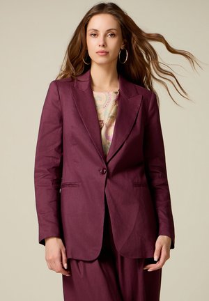 Vrouw met lang bruin haar, grote oorringen, een gebloemde blouse en een dichtgeknoopte bordeauxrode colbert en bijpassende broek.