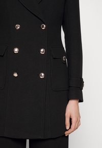 Manteau noir à double boutonnage avec des boutons dorés, coupe ajustée, deux poches à rabat et tissu structuré, présentant des coutures détaillées et des poignets.