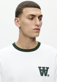 White sweatshirt with green ribbed collar and embroidered logo on the left chest. Smooth texture and relaxed fit.