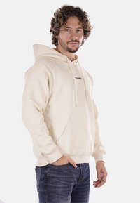 Felpa beige realizzata in tessuto morbido con tasca a marsupio, dotata di cappuccio con coulisse e piccolo logo nero sul petto.