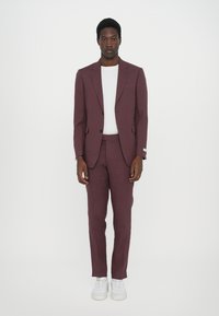 Bordeauxfarbener Anzug mit einem taillierten Blazer und passender Hose. Darunter ein weißes T-Shirt mit Rundhalsausschnitt. Modell trägt weiße Sneakers. Glatte Stoffstruktur.