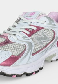 Λευκό, ροζ και γκρι παπούτσι τρεξίματος New Balance με διχτυωτές επενδύσεις, λευκά κορδόνια και υφή σόλας ορατή σε ανοιχτό φόντο.