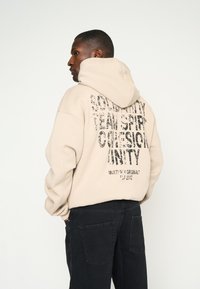 Sudadera beige con texto negro en la parte posterior que dice "ESPÍRITU DE SOLIDARIDAD EQUIPO COHESIÓN UNIDAD". Confeccionada en un material suave y de diseño holgado.