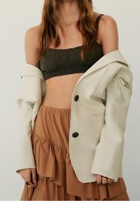 Blazer oversize beige clair avec boutons noirs, superposé à un crop top en tricot marron foncé et une jupe à volants superposés marron.