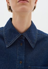 Denim shirt in dark blue with a classic collar, button closure, and visible stitching. Features a chest pocket and a textured fabric.