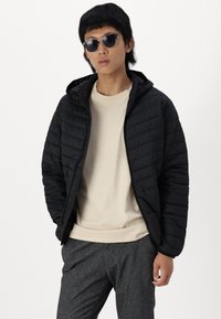 Schwarze gesteppte Jacke mit Kapuze, Reißverschluss vorne, kombiniert mit einem hellbeigen Sweatshirt und dunkelgrauen Hosen. Das Model trägt dunkle Sonnenbrillen.