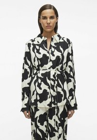 Camisa de patrón en blanco y negro con cuello, cierre de botones y cintura ceñida. Las mangas largas cuentan con aberturas laterales y puños ajustados.