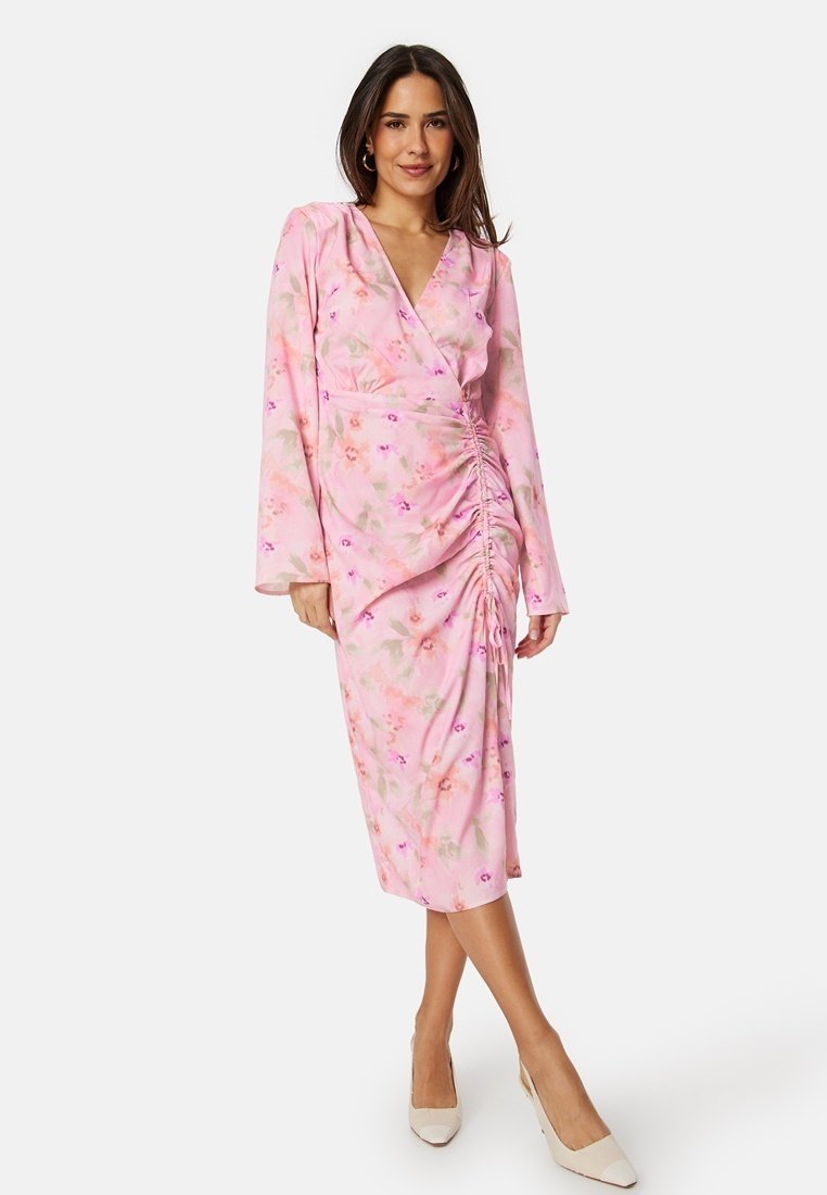 Rosa blommig omlottklänning med långa ärmar, rynkad sidodetalj och medellång design, tillverkad av lättviktsmaterial. Bärs med beige klackar.