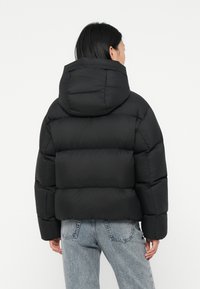 Svart puffjacka med huva, med kviltade sektioner och avslappnad passform. Bärs över ljusblå jeans, tyget verkar slätt och glänsande.
