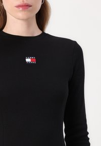 Fekete bordázott, hosszú ujjú felső, amelyen a mellkasán egy kis piros, fehér és kék hímzett logó olvasható: "TOMMY JEANS".
