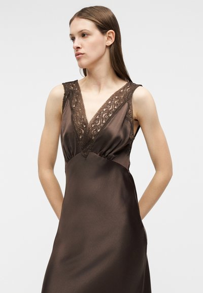 Νεαρή γυναίκα με αμάνικο σκουρό καφέ σατέν φόρεμα με δαντελένιο V-λαιμόκοψη, που κοιτάζει προς τα πλάγια με φόντο ένα απλό ανοιχτόχρωμο φόντο.