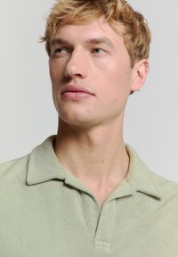 Lichtgroene polo met een gestructureerde stof, voorzien van een puntige kraag en een knopenlijst. De stiksels zijn zichtbaar en benadrukken het casual ontwerp.