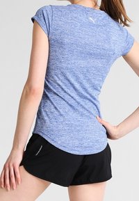 Mujer vestida con una camiseta atlética de manga corta azul y pantalones cortos negros, de pie con una mano en la cadera y la otra descansando a lo largo del cuerpo.