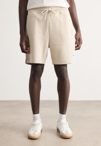 Shorts côtelés beiges avec taille élastique et cordon de serrage ajustable. Associés à des baskets blanches et des chaussettes mi-mollet, se tenant à l'intérieur.