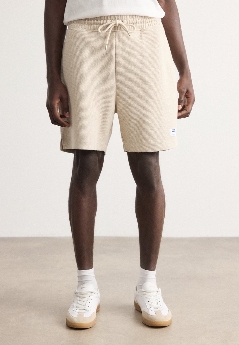 Shorts côtelés beiges avec taille élastique et cordon de serrage ajustable. Associés à des baskets blanches et des chaussettes mi-mollet, se tenant à l'intérieur.