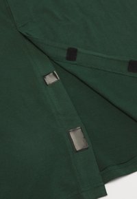 Tmavě zelené tkaninové tričko s šedými suchými zipy na lemu, hladkou texturou a jednoduchým designem bez dalších vzorů nebo akcentů.