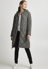 Lange, met kap, grijze gewatteerde pufferjas met een rits aan de voorkant, een ruitpatroon en zijzakken, gecombineerd met zwarte broek en witte sneakers.