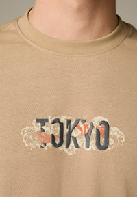 Camiseta de manga corta beige con cuello redondo que presenta el texto "TOKIO" en azul y rojo, adornado con patrones de olas en colores claros.