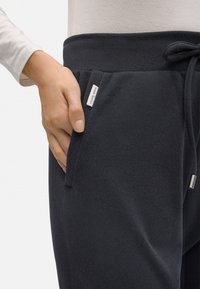 Pantalones de chándal gris oscuro con tejido texturizado, bolsillo lateral con etiqueta de logo y cintura con cordón. Material suave y corte relajado.