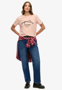 Camiseta rosa claro con estampado negro, camisa de cuadros roja atada a la cintura, jeans de mezclilla azul y zapatos negros, de pie frente a un fondo blanco.